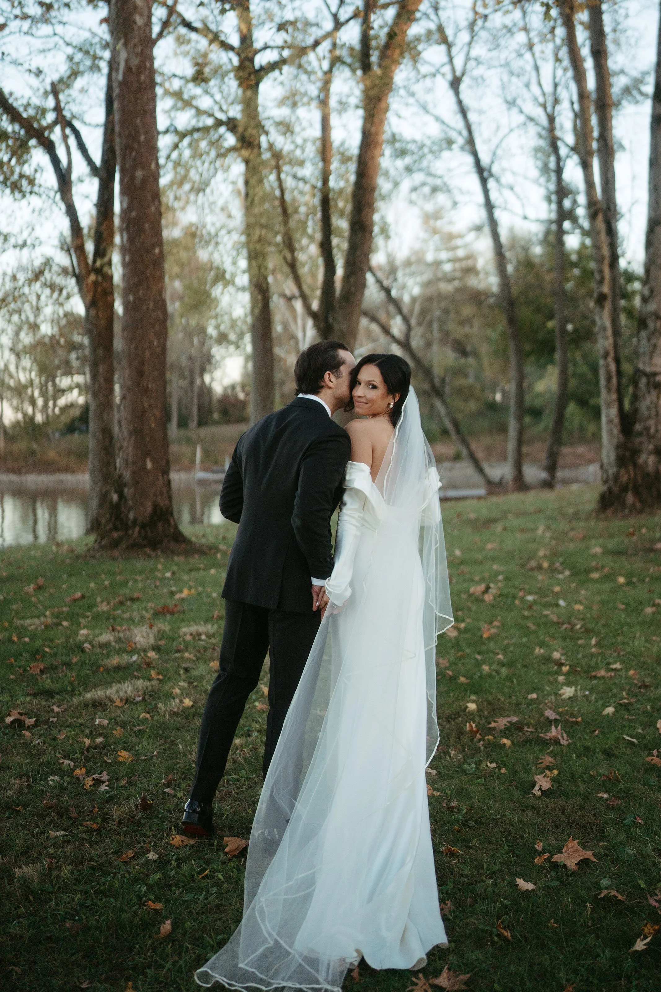 A bride and groom holding hands outdoors near a lake, surrounded by tall trees, on their wedding day.