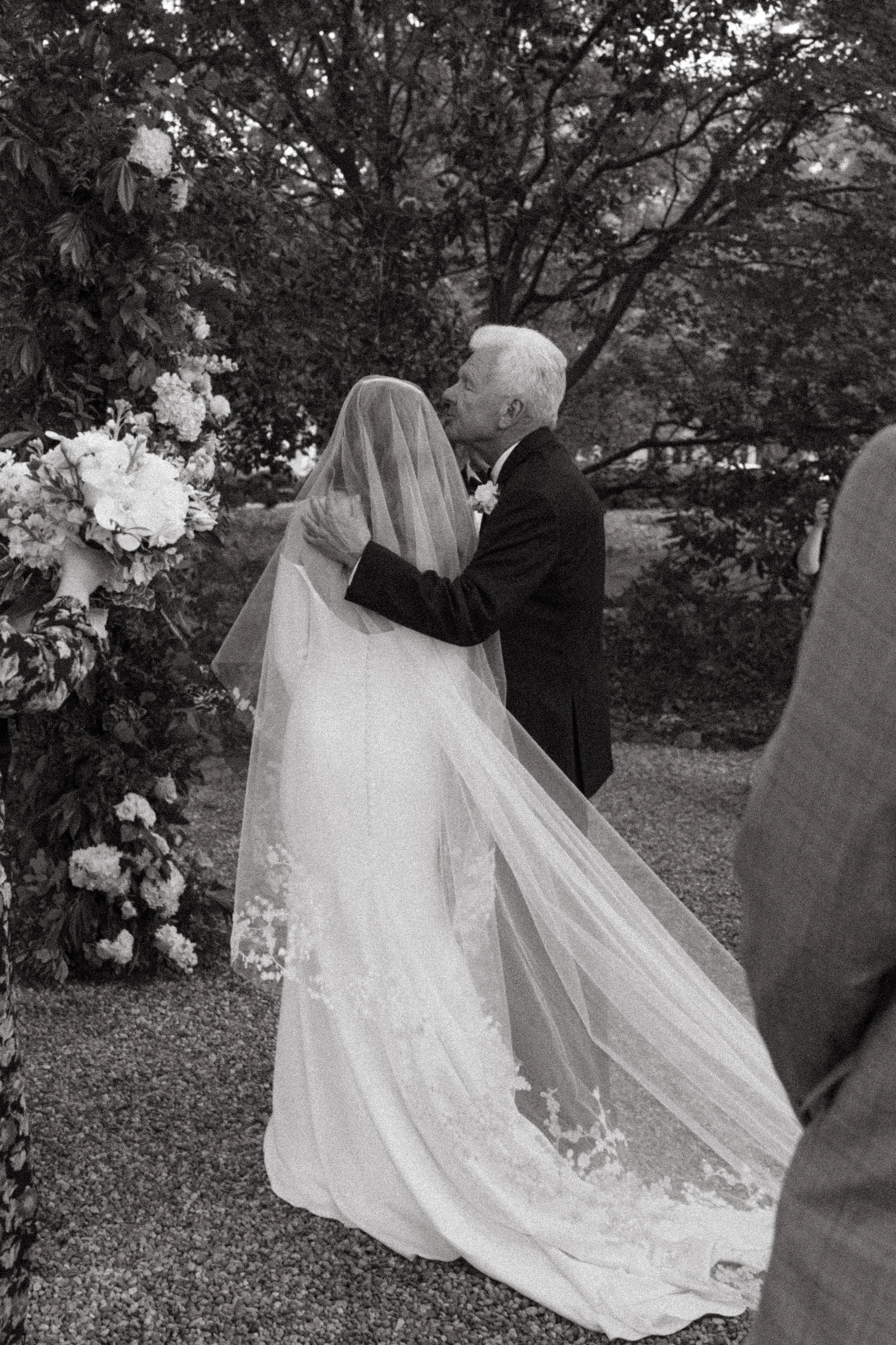 A black-and-white photo of a wedding ceremony outdoors, showing a bride in a wedding gown and veil being embraced and kissed by an older man in a tuxedo, with a floral arch and trees in the background.