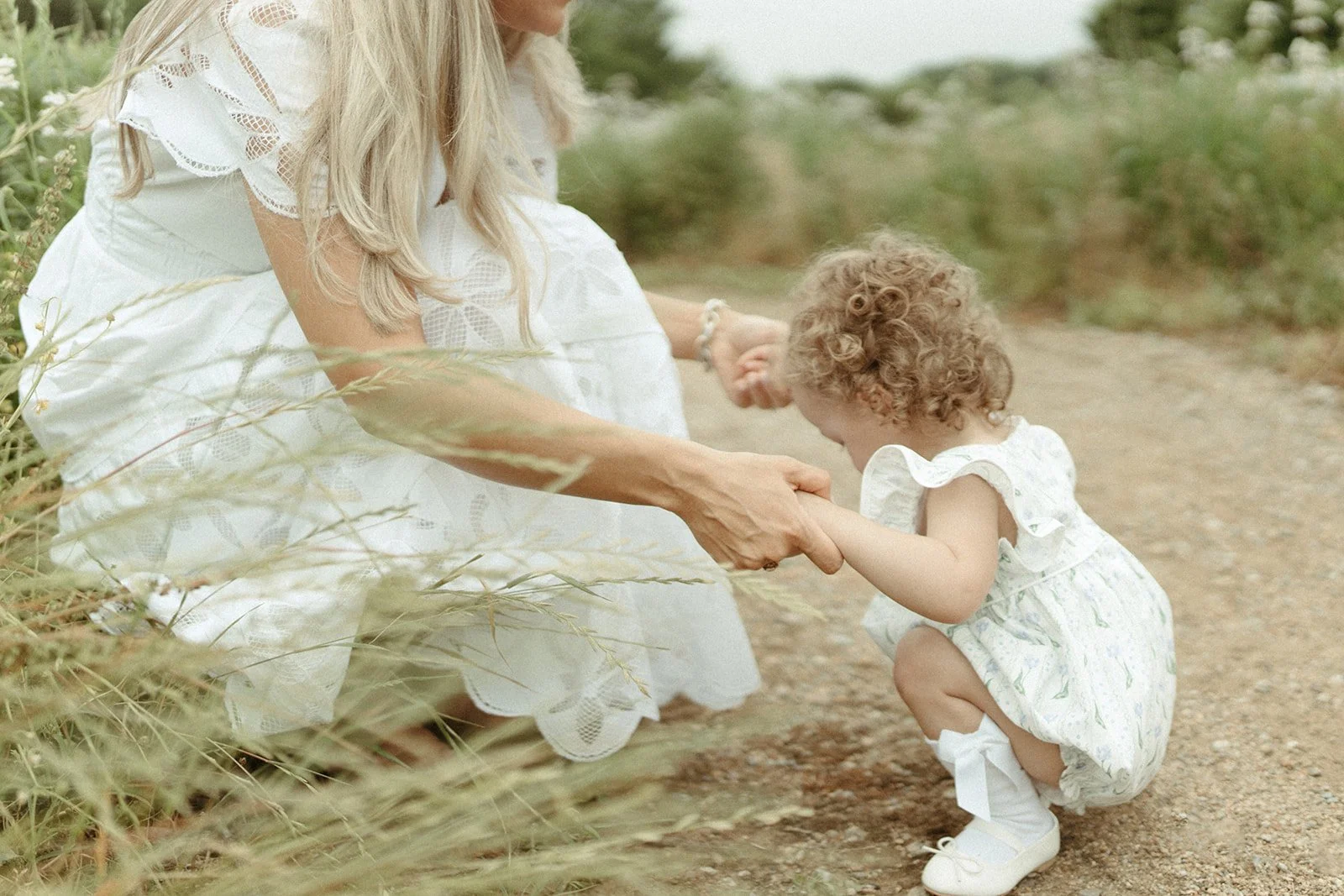 A woman in a white dress kneeling on the ground and holding hands with a young girl in a white dress with floral patterns, who is squatting and holding her hand, on a dirt path surrounded by green vegetation.