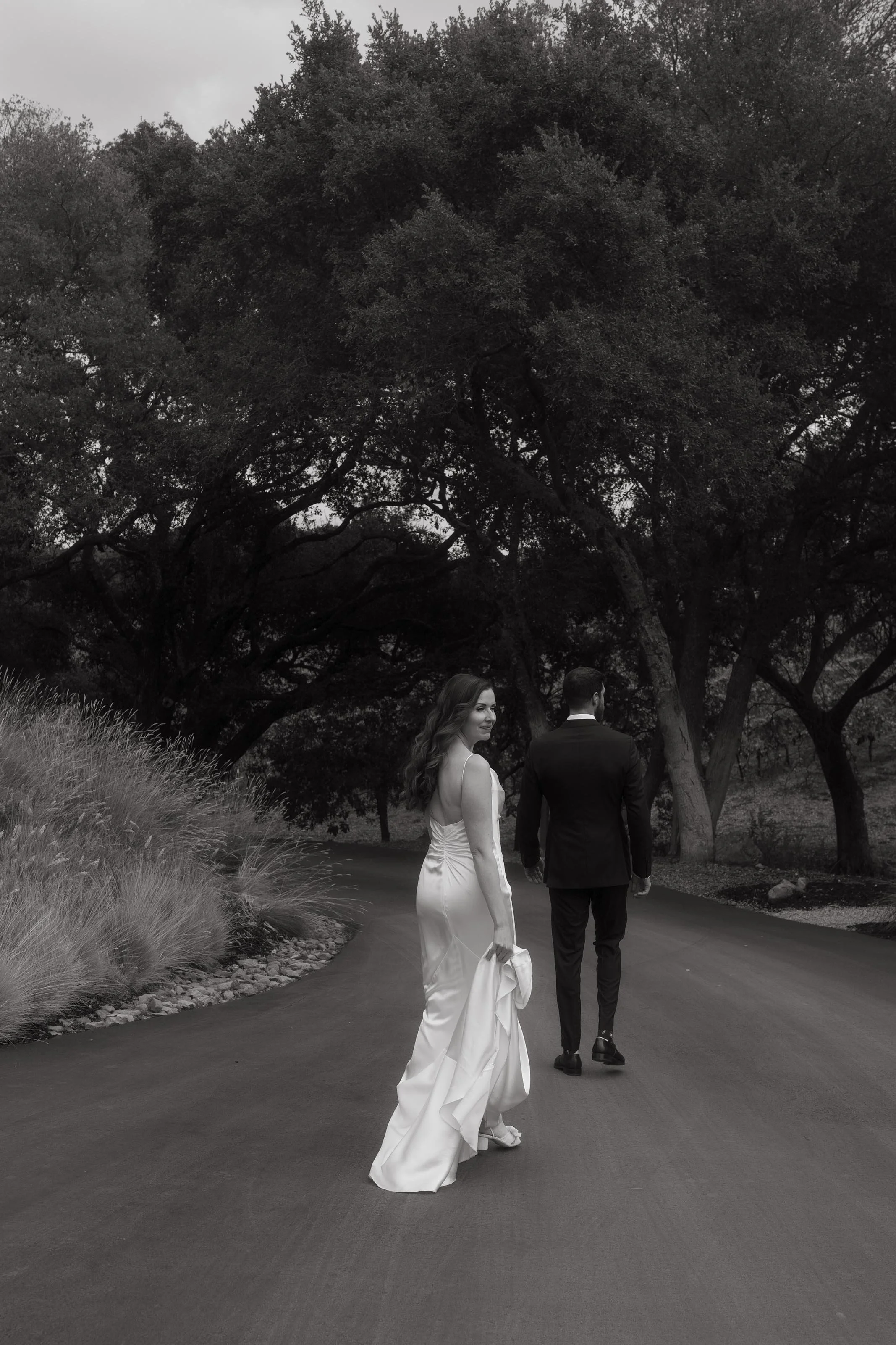 A bride and groom walk along a winding path in a park, with the bride turning back and smiling, surrounded by trees and tall grasses, in black and white.