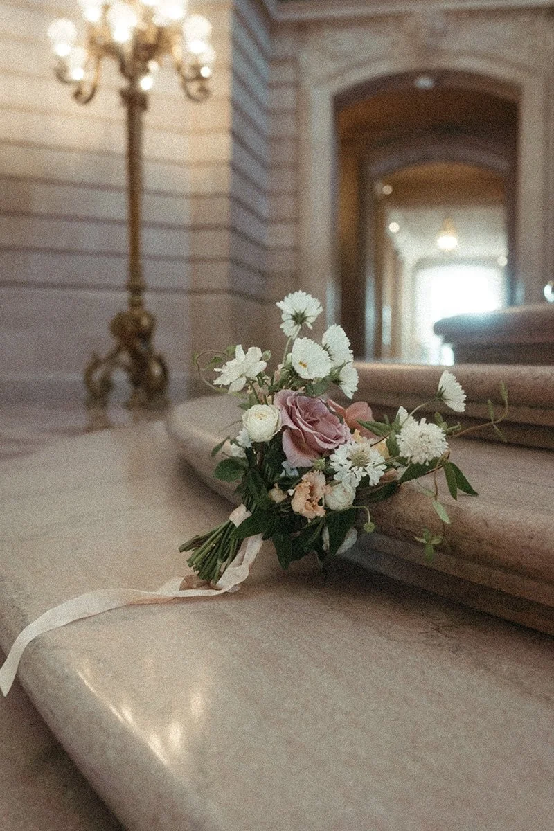 A bridal bouquet with pink and white flowers resting on a beige fabric surface in an elegant room with a chandelier and wooden arches.