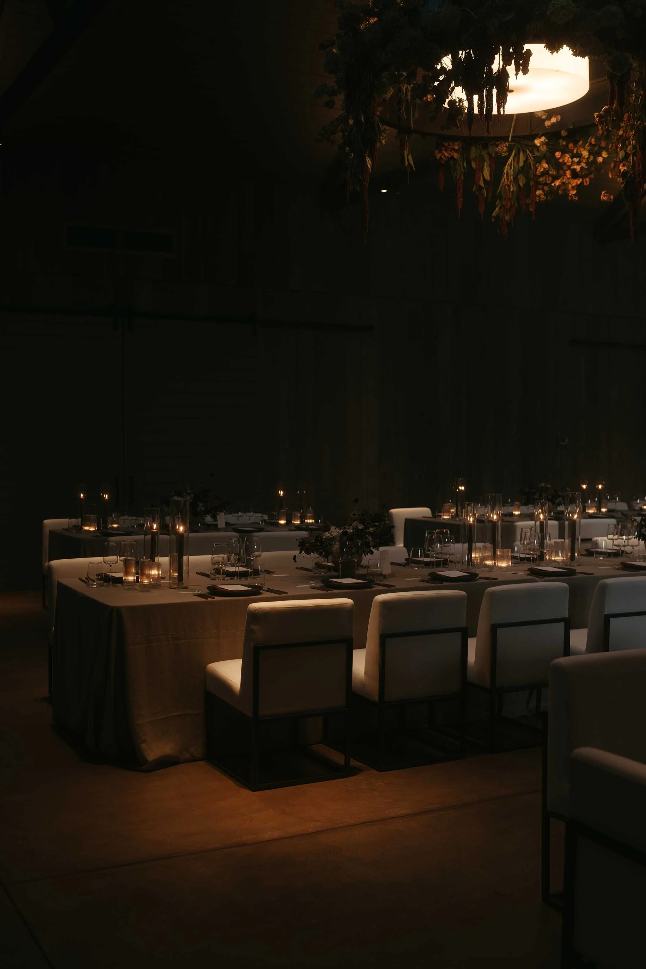 A dimly lit indoor dining setup with a long table, white chairs, and candlelit centerpieces. Overhead, a floral and greenery arrangement decorates a circular light fixture.