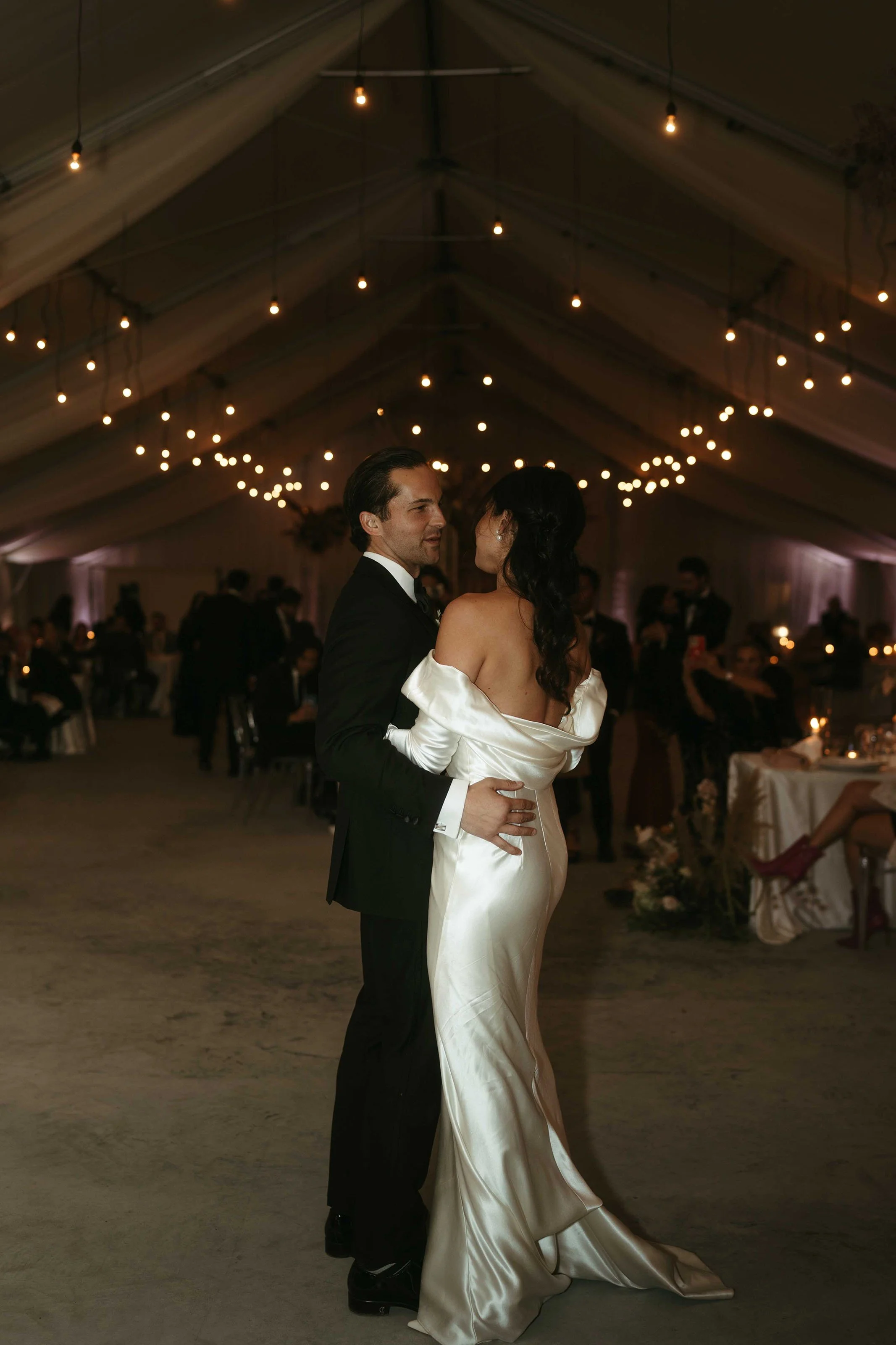 A couple dancing at their wedding reception inside a decorated tent with hanging string lights, surrounded by seated guests.
