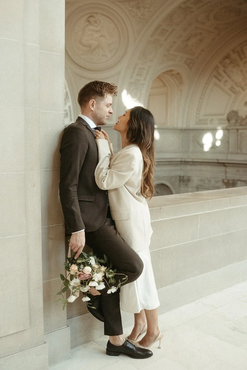 A romantic couple in formal attire stands close together in an elegant, historic building. The man is leaning against a wall, holding a bouquet of flowers, as he looks at the woman, who is touching his shirt collar and gazing at him. The woman wears 