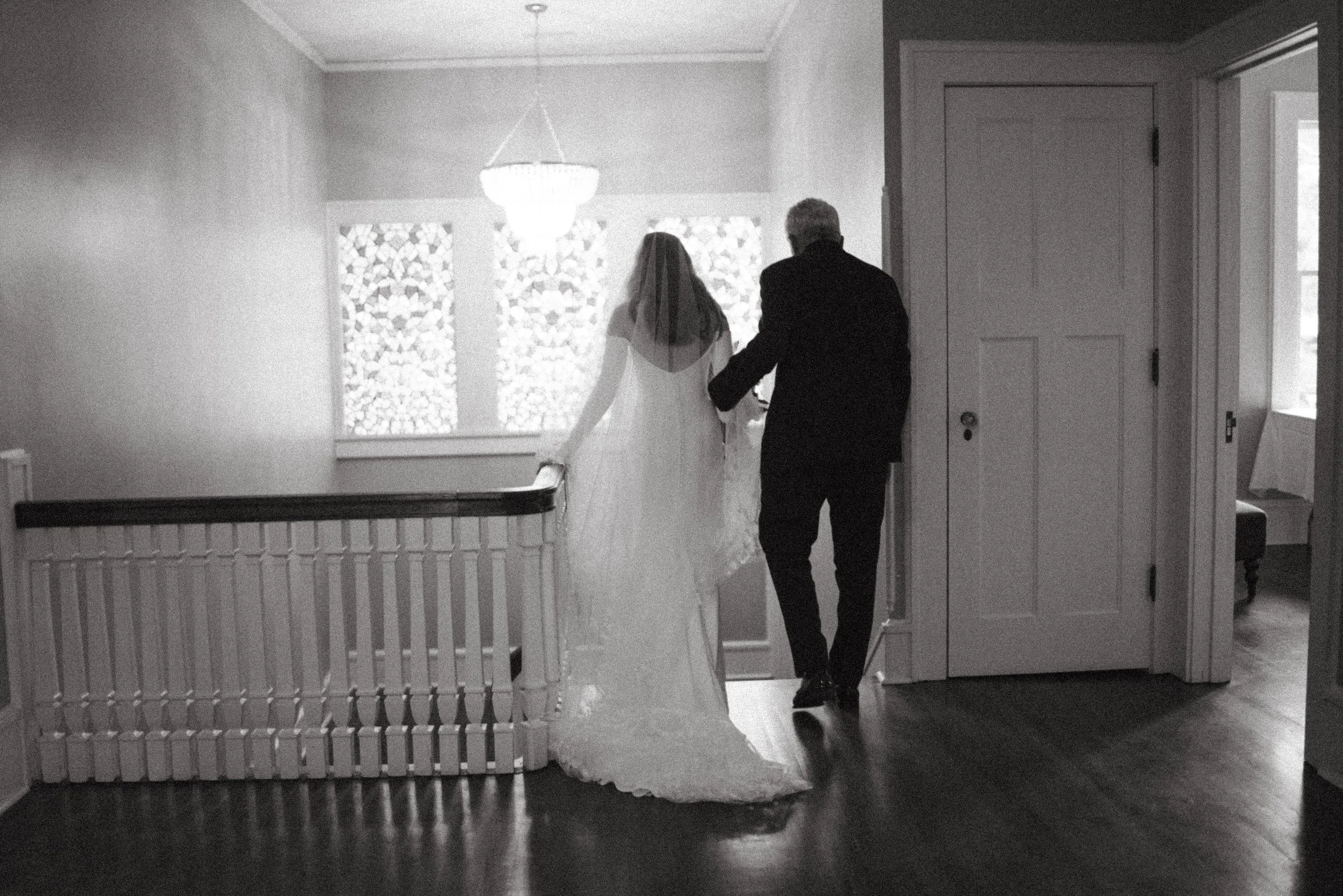 A bride in a wedding dress walking with an older man in a suit down a hallway inside a house.
