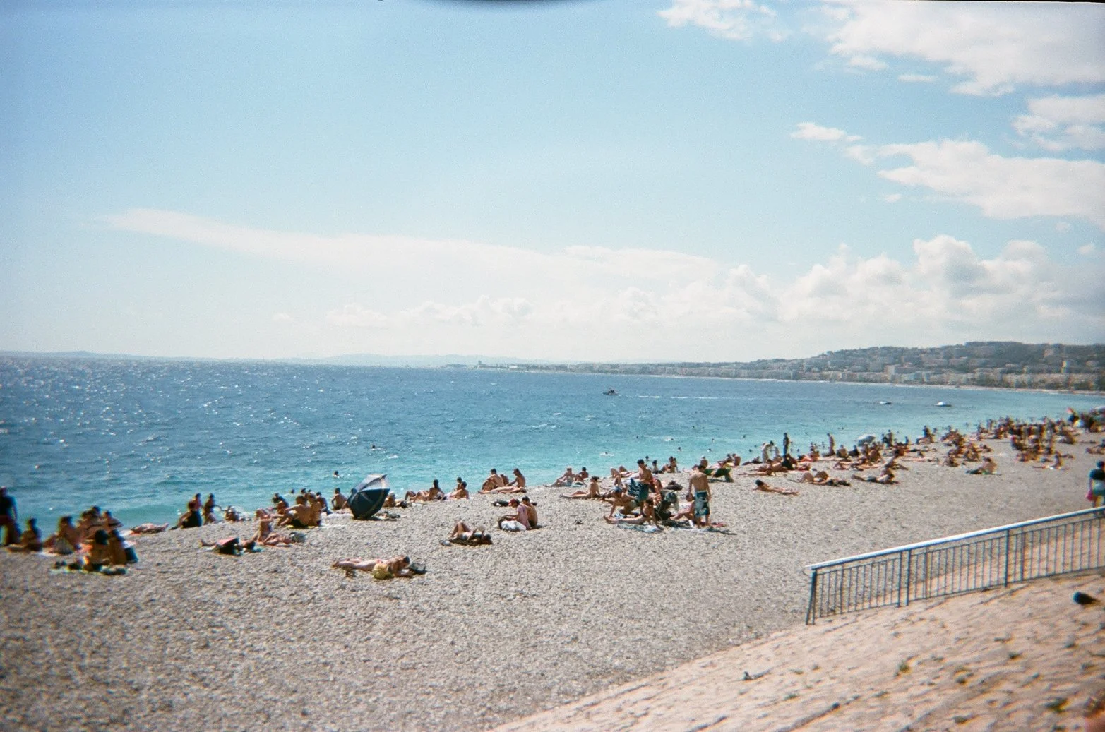 Beach scene with many people sunbathing, swimming, and relaxing on a pebbled shoreline, under a partly cloudy sky.