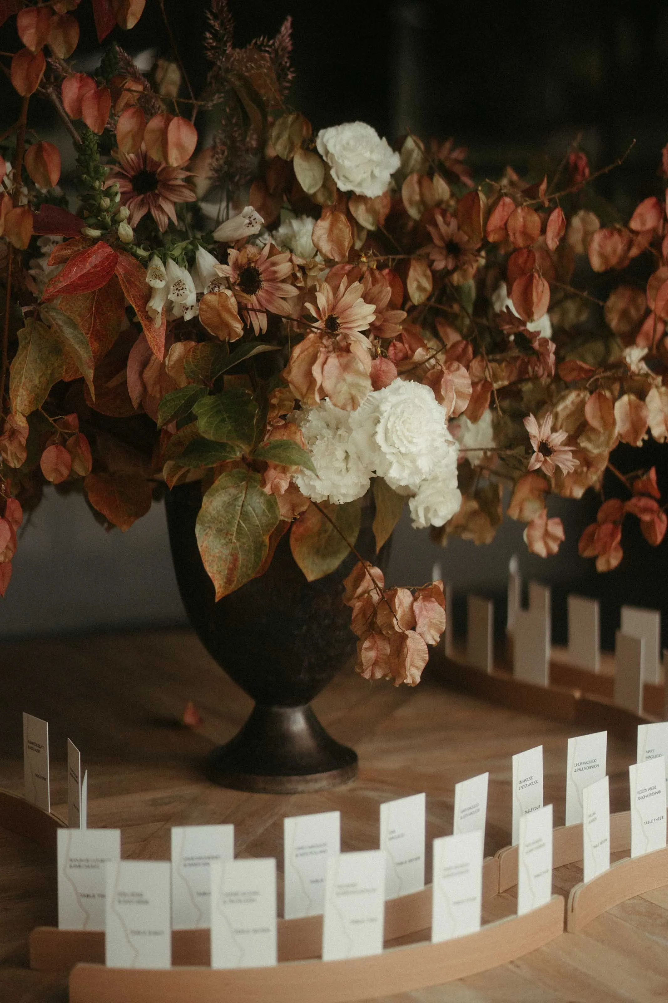A large black vase filled with autumn-colored flowers and dried leaves, placed on a wooden surface, with white paper tags arranged in a curved line in front of it.