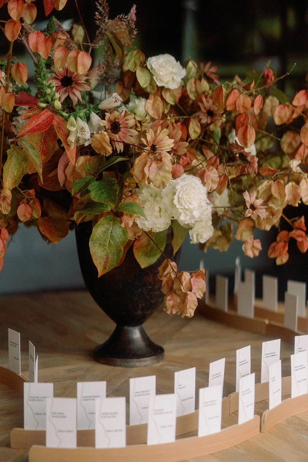 A dark vase holding a large arrangement of dried flowers and leaves on a wooden table, with small white cards in front.
