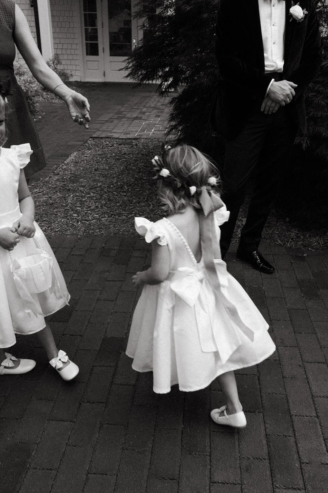 A young girl in a white dress with floral decorations and shoes with bows, standing on a brick walkway outdoors, with other children and an adult nearby.