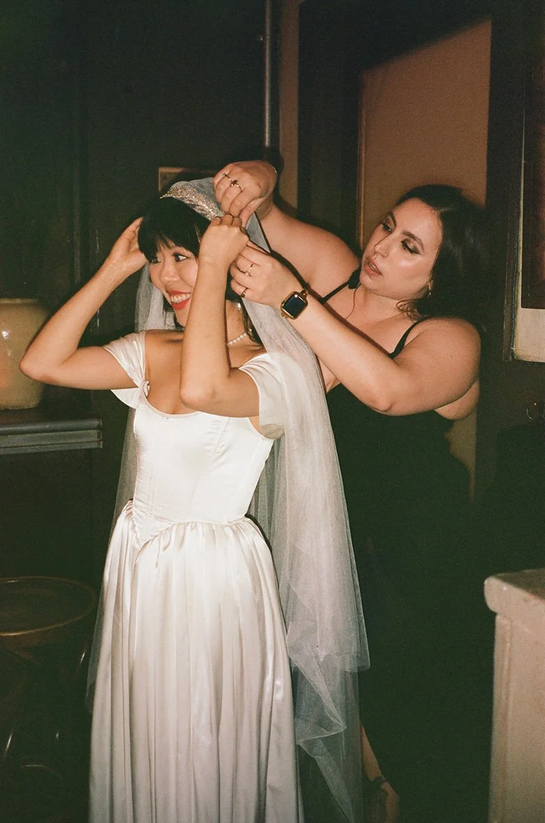 A woman in a white satin dress is getting her veil adjusted by another woman with a black dress, in a dimly lit indoor setting.