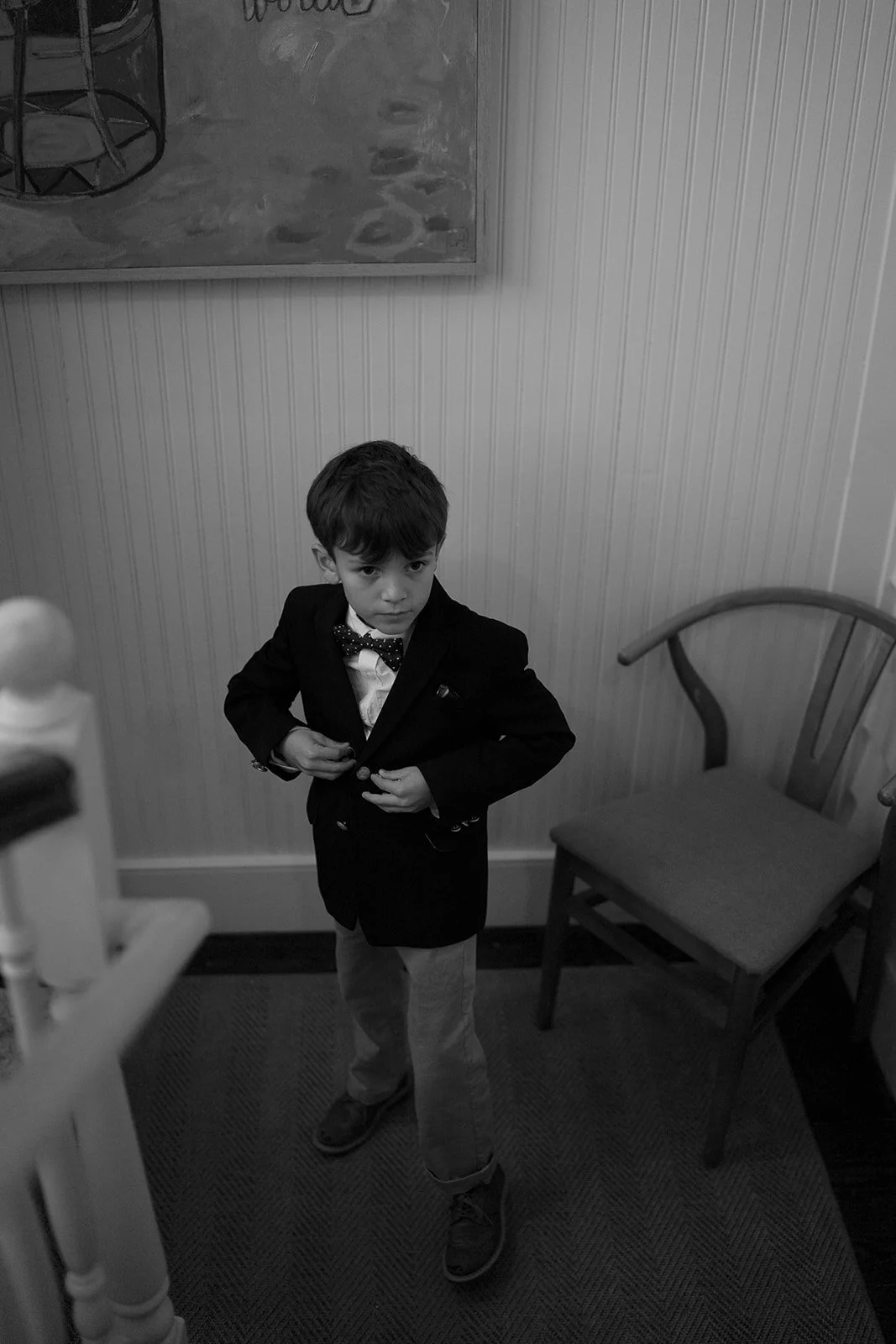 A young boy dressed in a black suit, white shirt, and polka dot bow tie standing in a room with a chair and wall paneling, adjusting his jacket and looking at the camera.