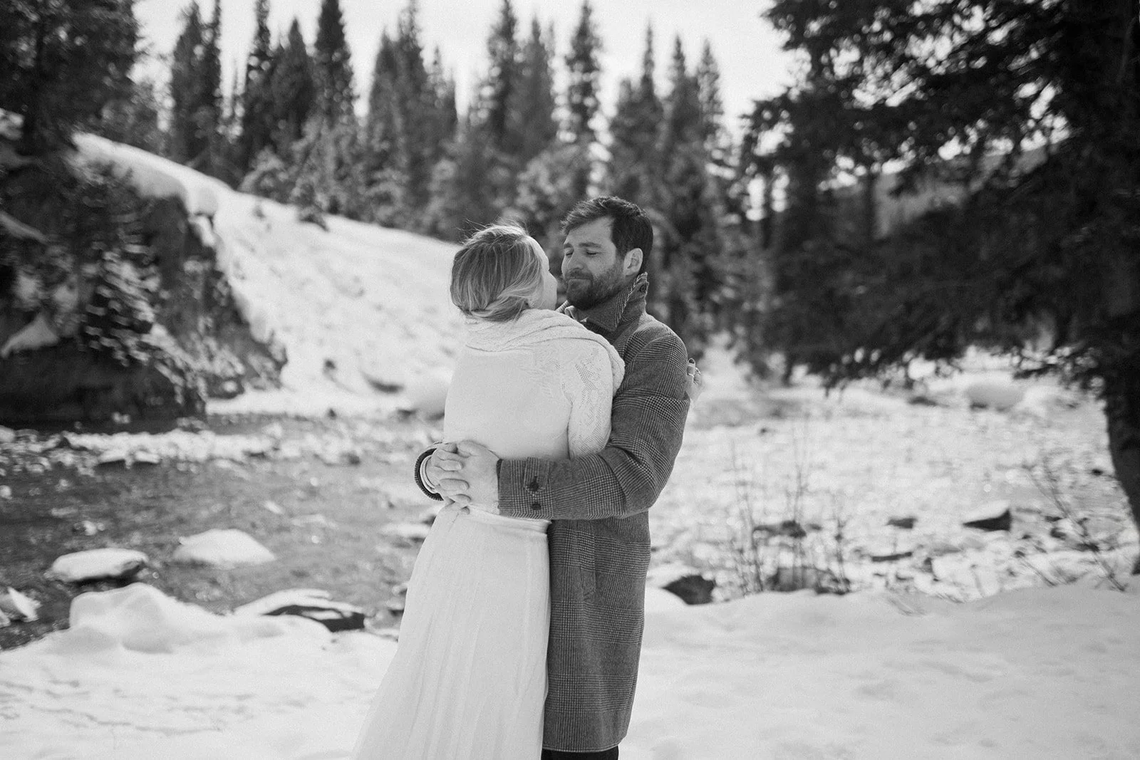 A black and white photo of a couple embracing outdoors in a snowy landscape with trees in the background.