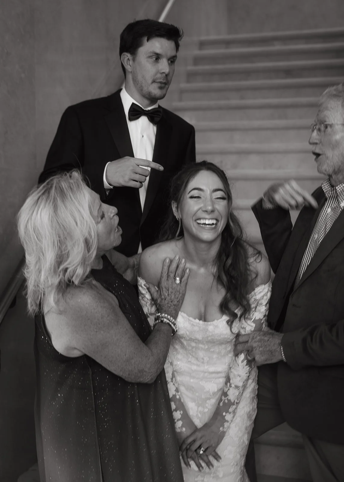 A joyful woman in a wedding dress laughing while surrounded by her family and friends at her wedding celebration, with some family members touching her shoulders and arm as they converse.