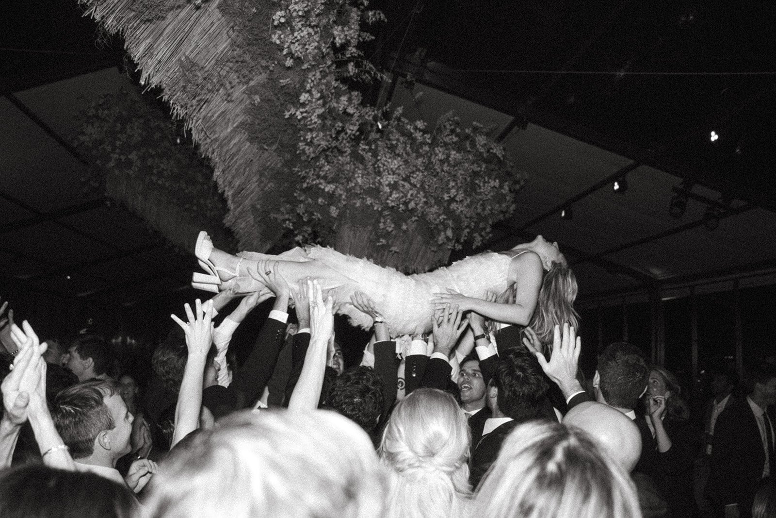 A group of people at a party or celebration lifting a woman in a wedding dress above their heads.