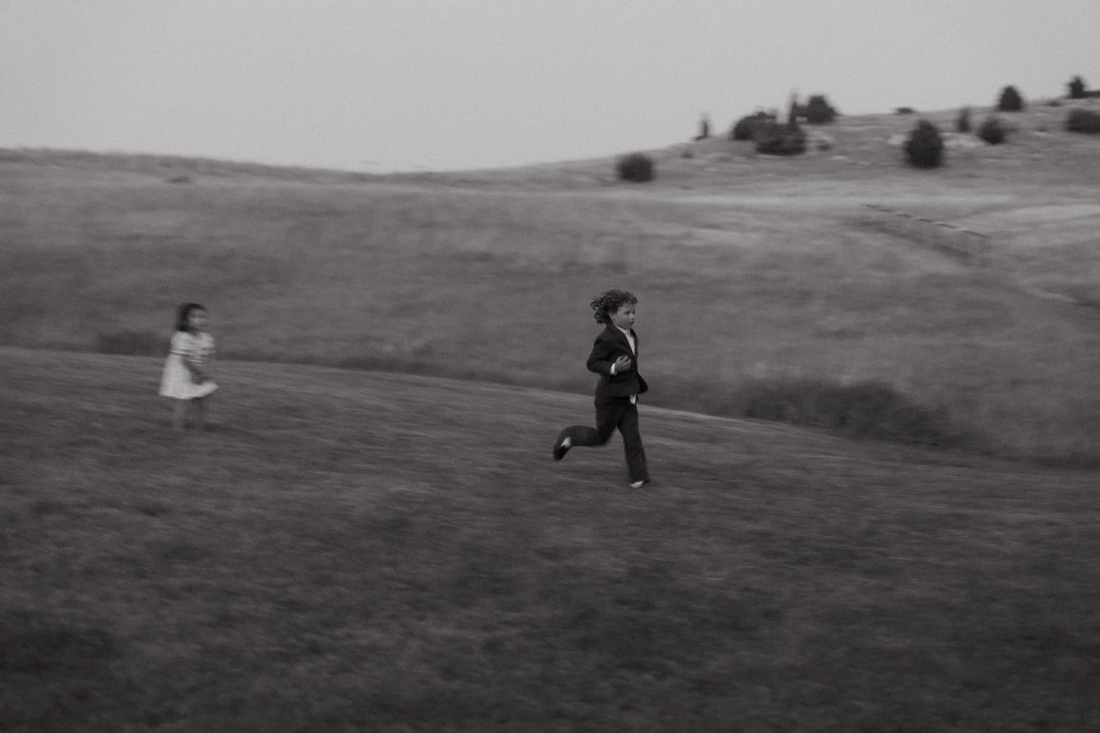 Black and white photo of a young girl and a young boy running in a grassy field with rolling hills and sparse trees in the background.