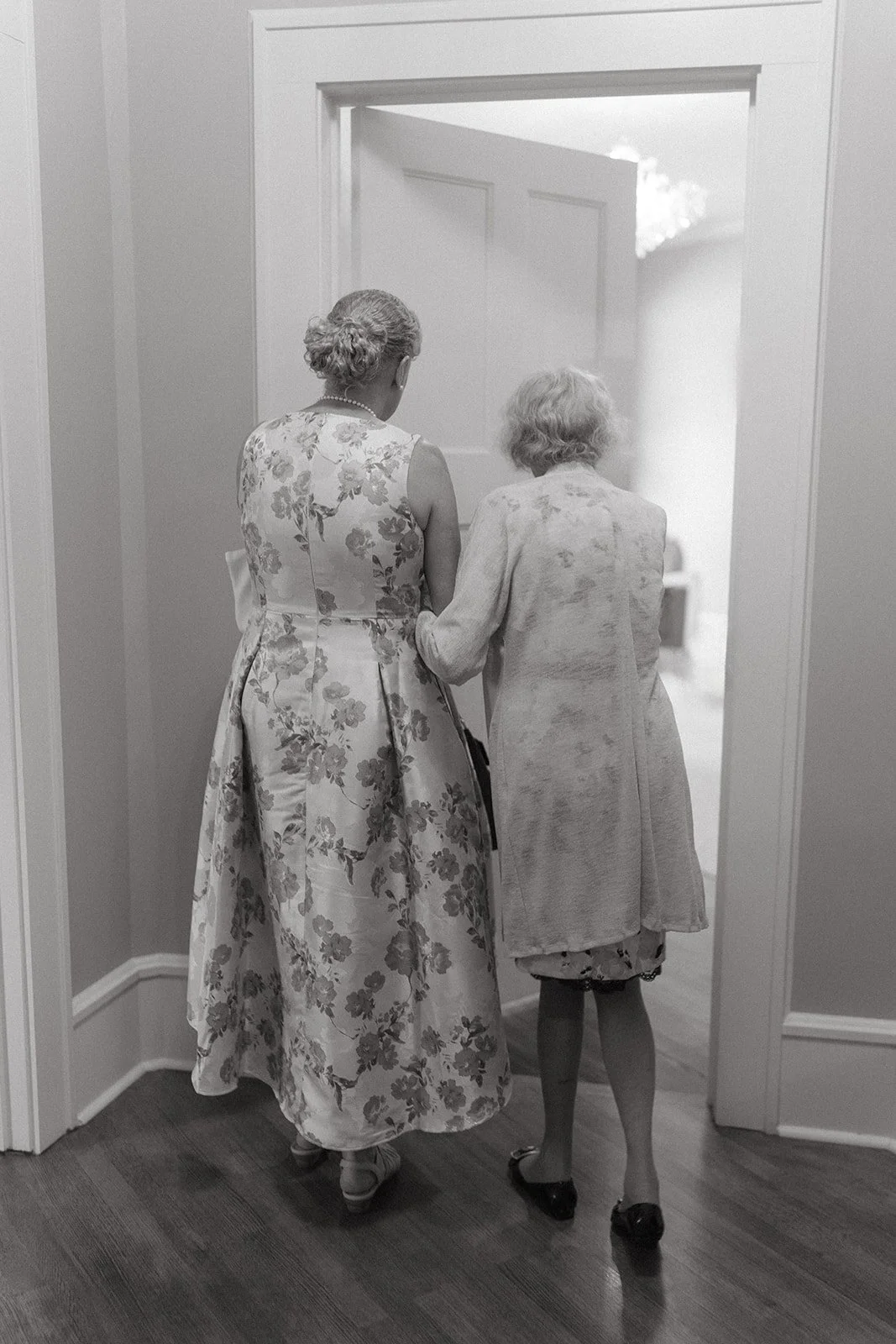 Two elderly women with gray hair, one in a floral dress and the other in a coat, walking arm in arm through a doorway in a domestic setting.