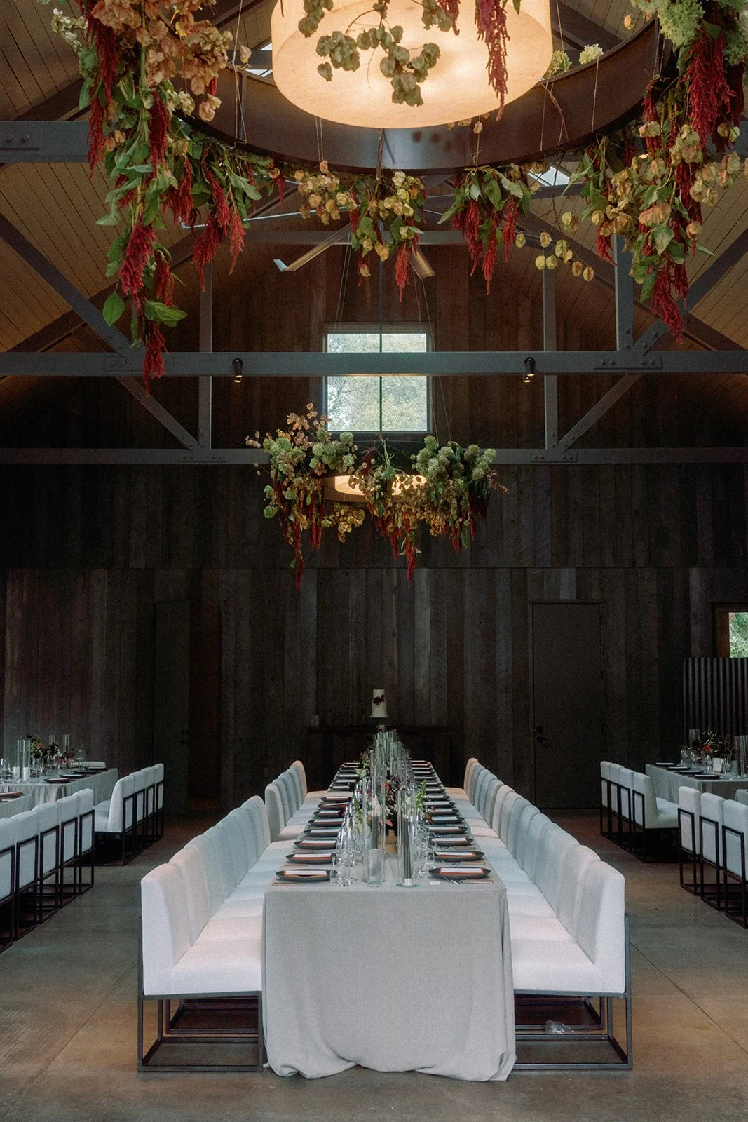 Elegant indoor dining area with a long, white-draped table set with glassware, plates, and silverware, surrounded by white chairs with black frames. The ceiling has rustic wooden panels and hanging floral arrangements with green, white, and red flowe