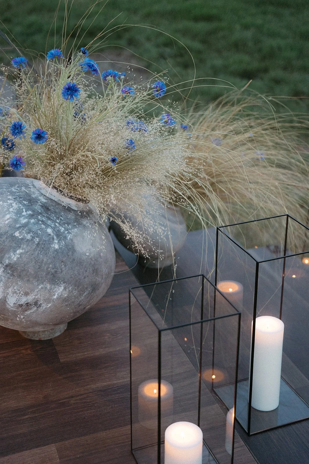 A decorative indoor or outdoor setup featuring a large, weathered, round gray vase filled with a bouquet of dried grass and vibrant blue flowers, next to two glass lanterns with white candles inside on a wooden surface, with a blurred green backgroun