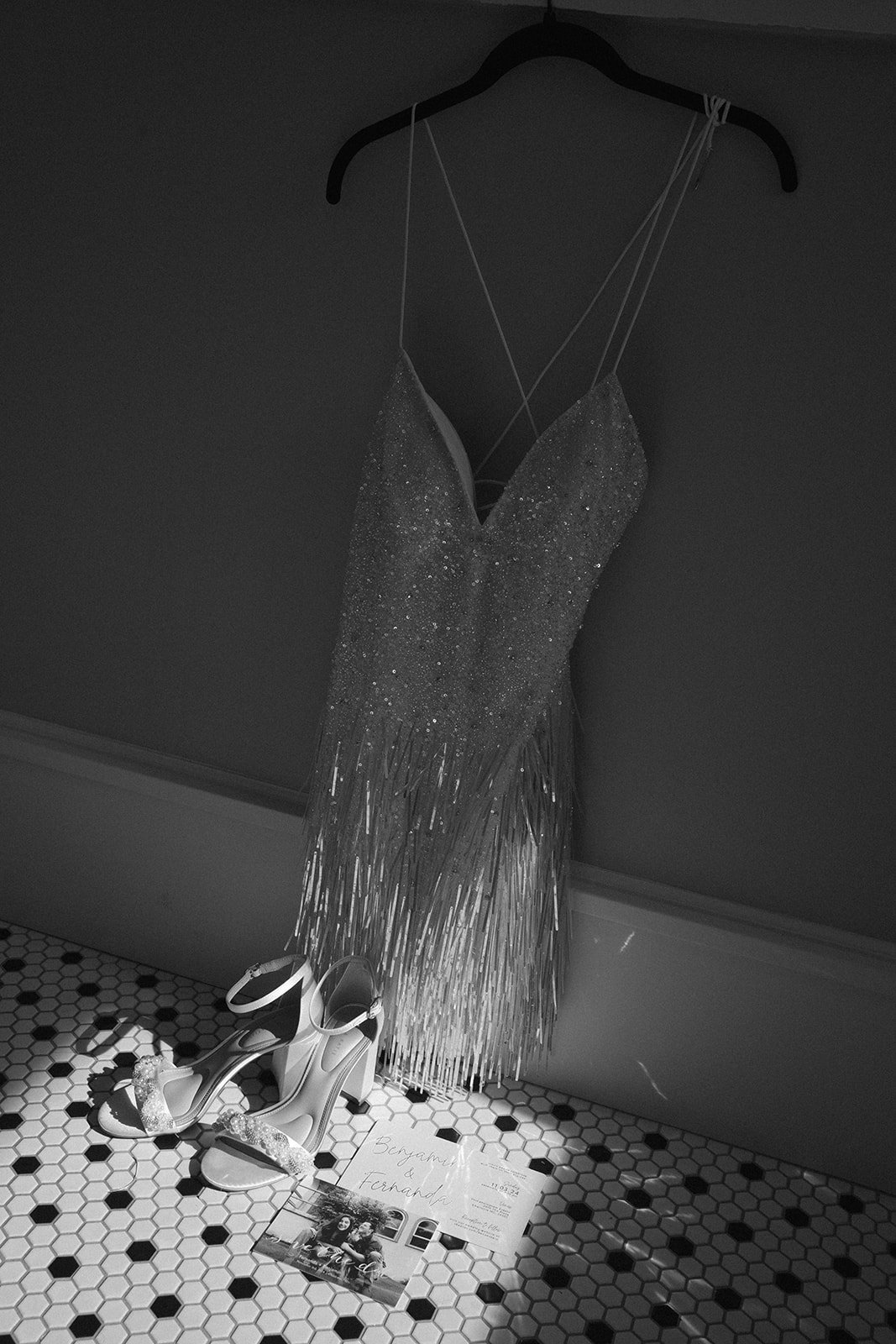 A sparkly, fringed evening dress hanging on a black hanger, with a pair of high-heeled sandals and a wedding invitation on a black-and-white hexagonal-tile floor.