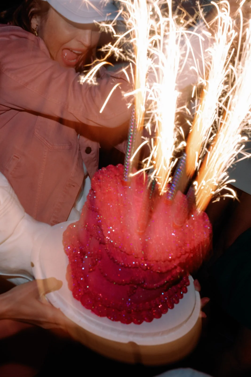 A woman in a pink jacket and sunglasses celebrates with a bright pink cake decorated with glitter and sparklers, exuding joy.