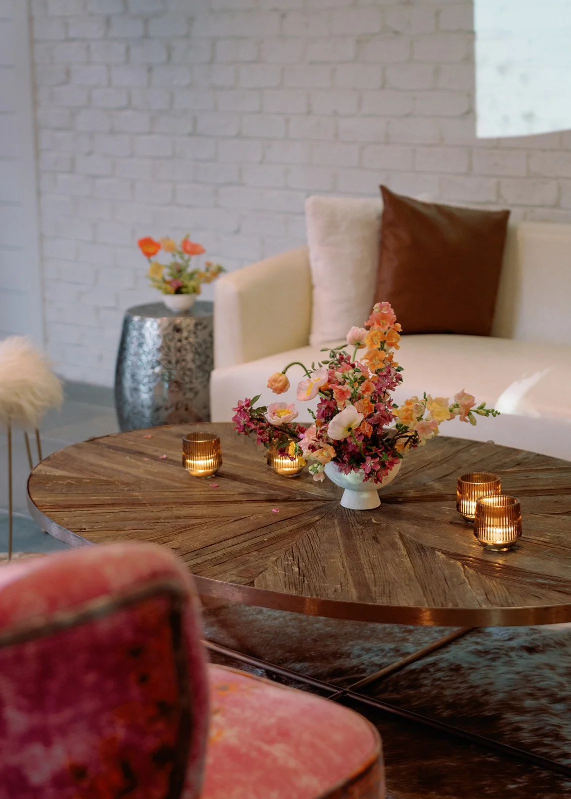A living room with a round wooden coffee table decorated with a vase of pink and yellow flowers and four small candle holders with candles. In the background, there's a white sofa with a brown pillow, and a silver side table with a flower arrangement