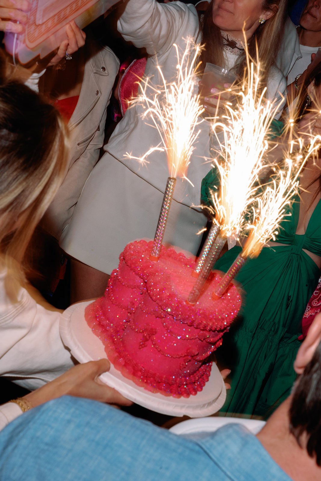 A birthday cake with pink frosting and sparkles, decorated with three sparklers that are lit and shooting sparks, surrounded by people celebrating.