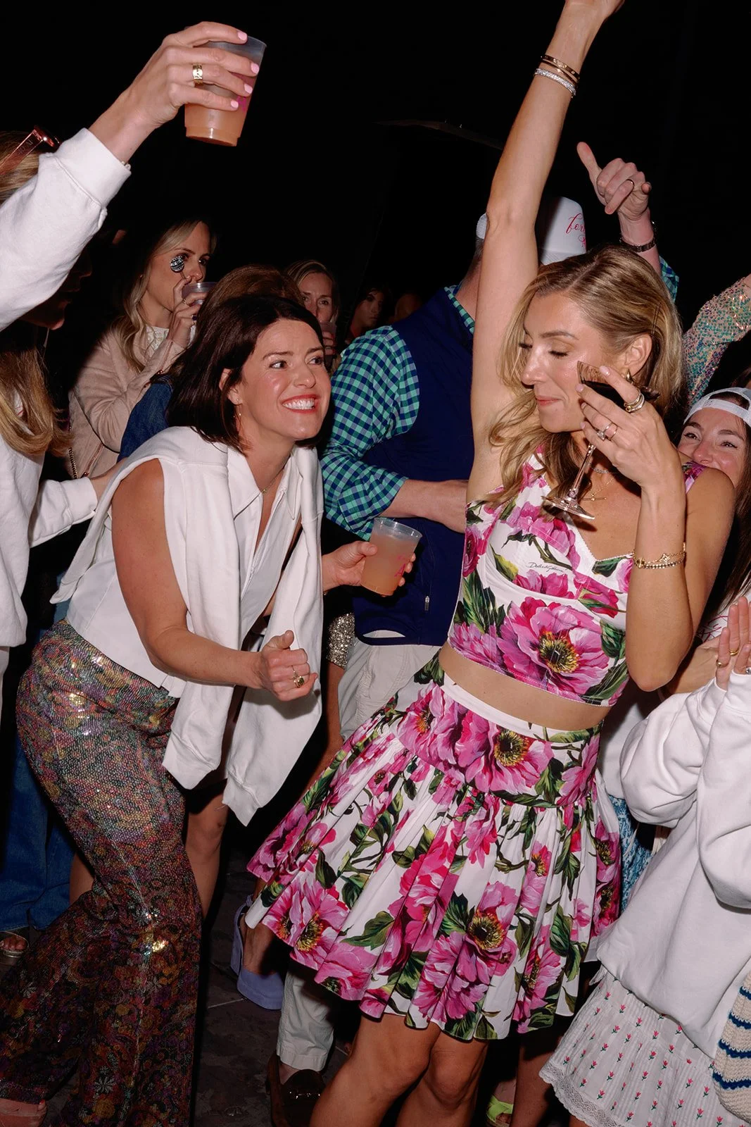 Women dancing and celebrating at a party with drinks, dressed in colorful outfits, with a dark background.