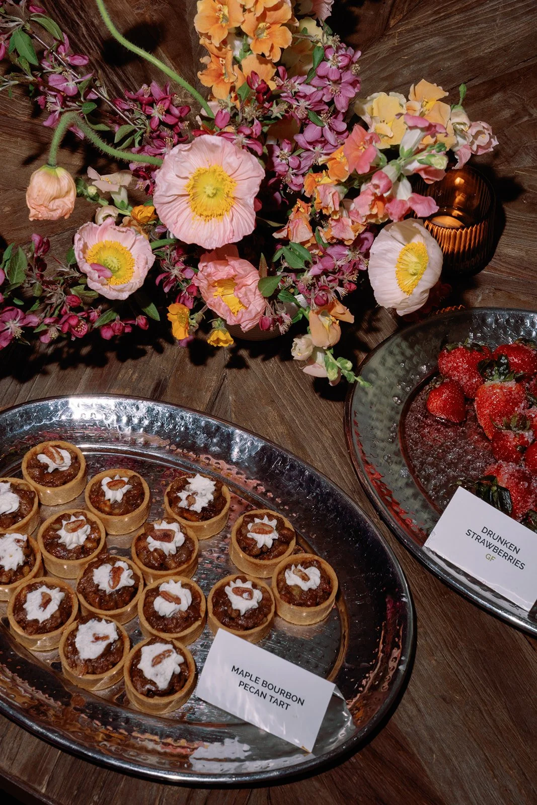 A floral arrangement of pink, orange, and purple flowers next to a plate of maple bourbon pecan tarts garnished with whipped cream and pecans, and a bowl of strawberries with a sign labeling it 'Drunken Strawberries'.