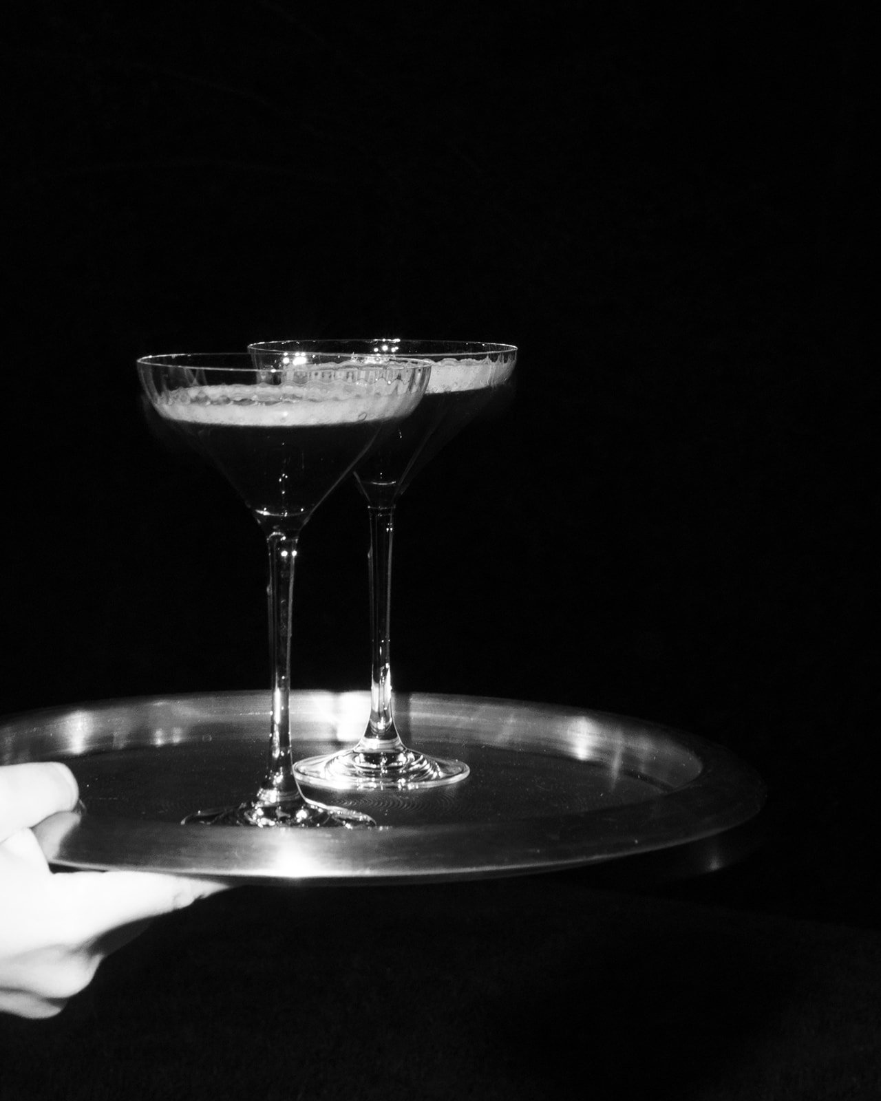 Two empty cocktail glasses on a silver tray held by a hand, against a dark background.