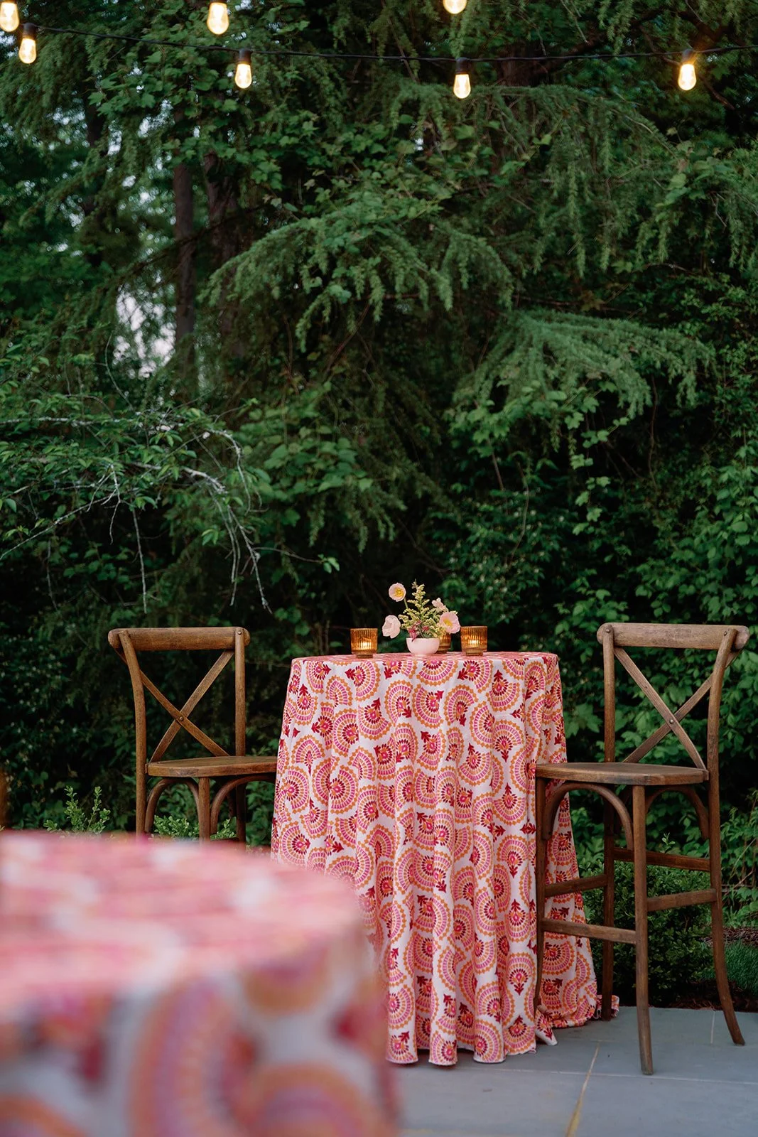 Outdoor dining setup with a round table covered with a colorful patterned tablecloth, flanked by two wooden chairs, situated among lush green trees, with string lights overhead and a small floral centerpiece on the table.