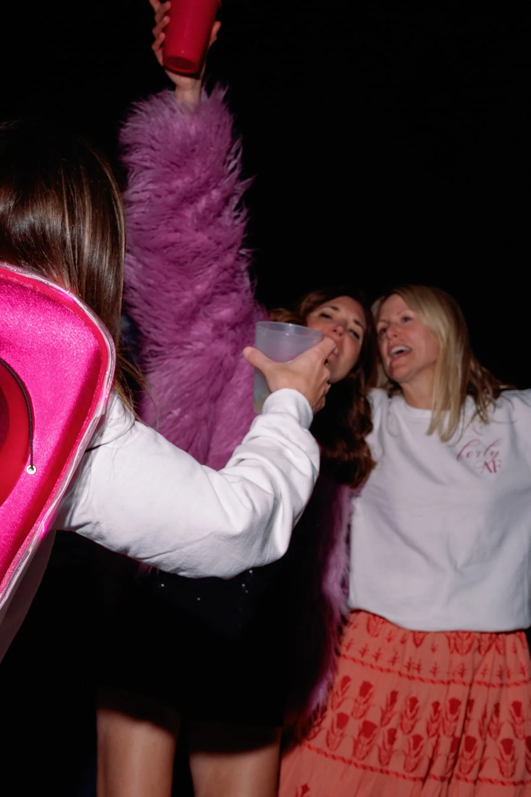 Group of people celebrating at a nighttime party, with one person pouring a drink into another's mouth, others laughing, and a person dressed in a pink furry costume.