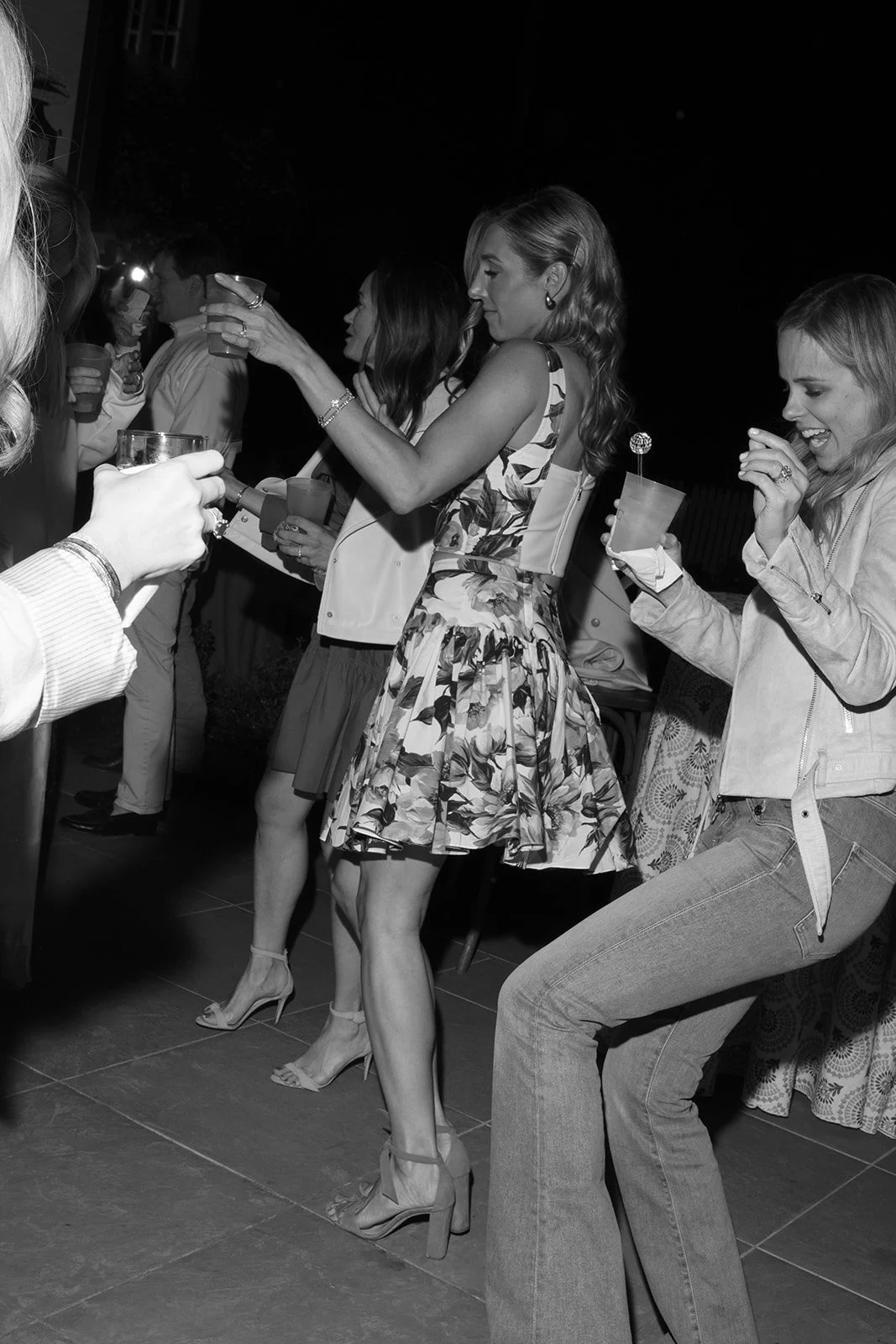People at a party holding drinks, dancing, and smiling, in an outdoor setting at night.