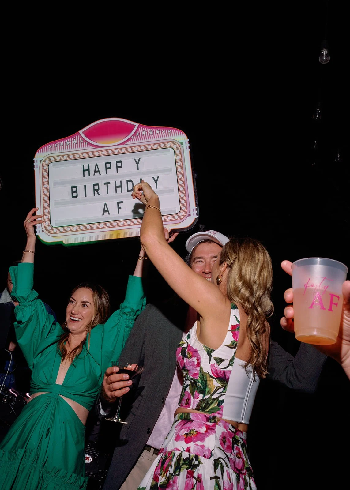 People celebrating at a party, holding a sign that reads 'Happy Birthday AF' and wearing festive attire.