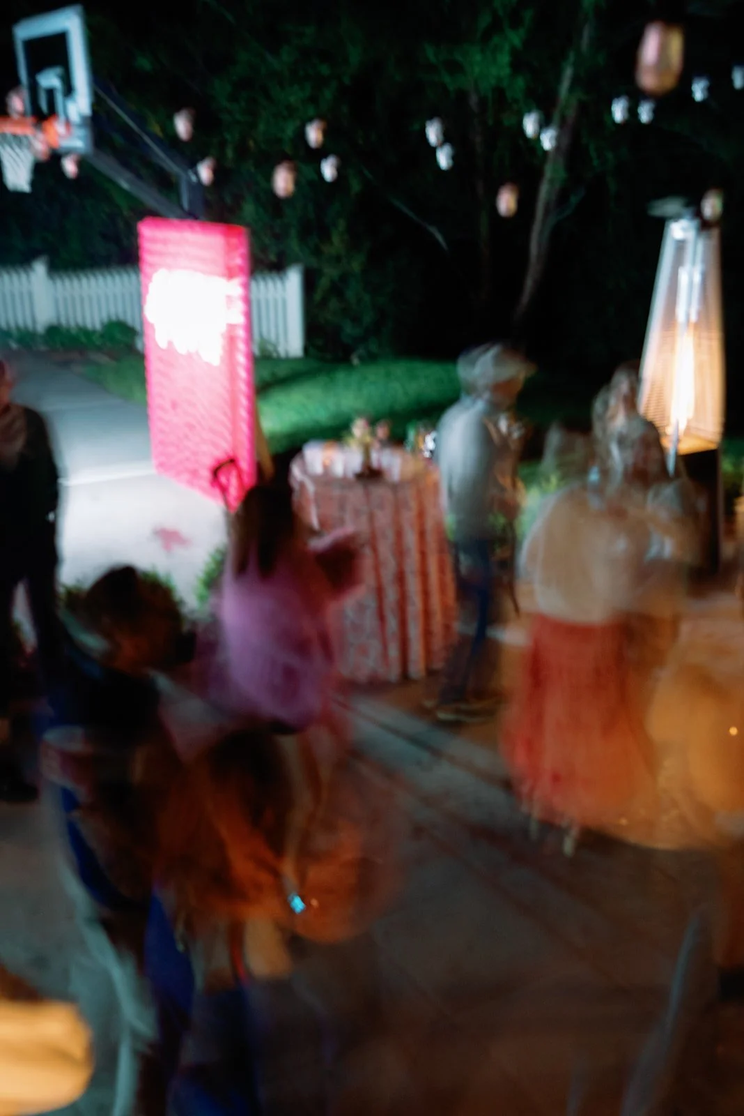 Nighttime outdoor gathering with blurry people, string lights, a pink sign, a table with decorations, and a basketball hoop in the background.