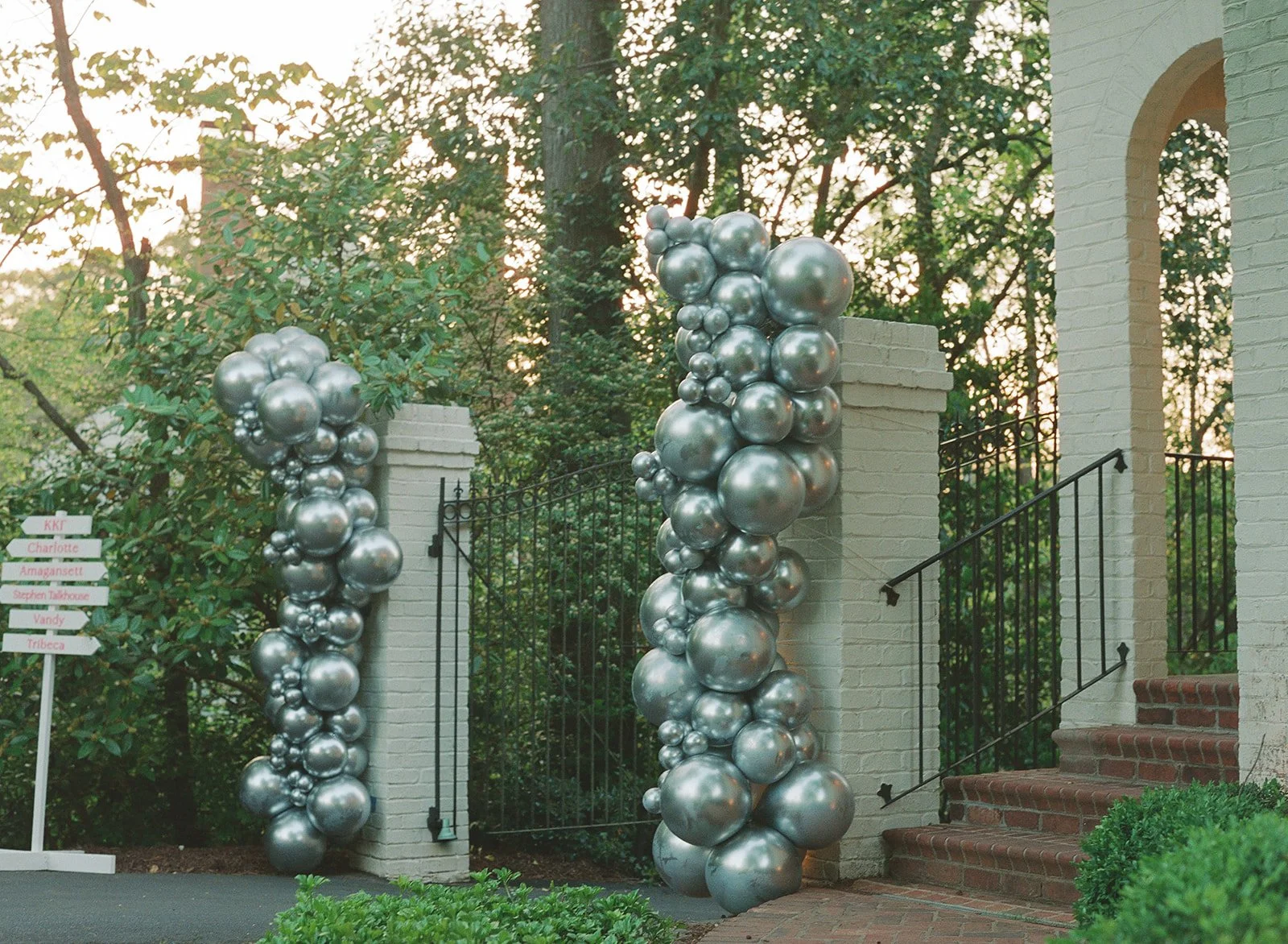 Decorative metallic balloon columns at the entrance of a house with stone steps and a white brick wall, surrounded by trees and greenery.