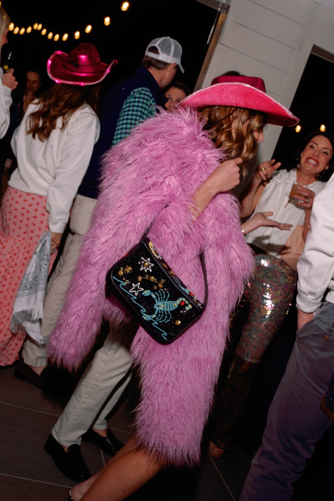 People celebrating at a party, with one woman in a pink faux fur coat and matching pink cowboy hat, holding a colorful bag with celestial and scorpion designs, and others in festive attire, including some women in polka dot skirts and cowboy hats.