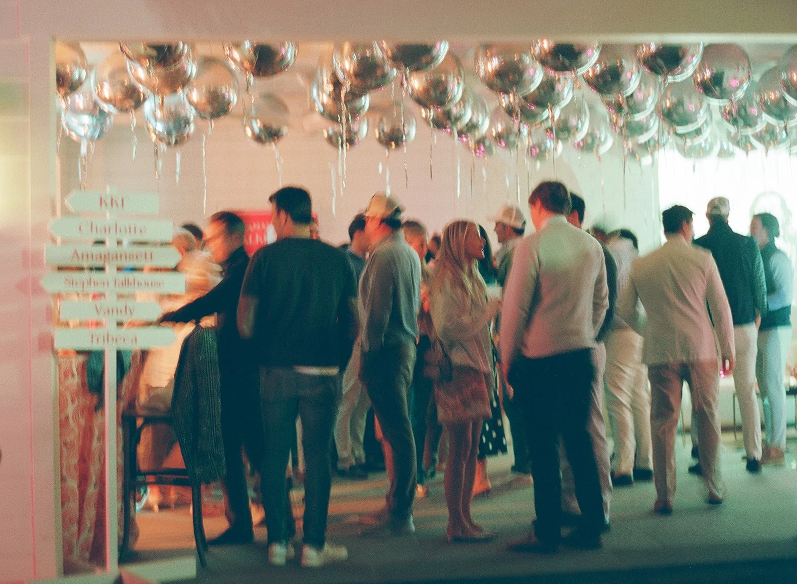 A group of people gathered in a room decorated with numerous silver balloons hanging from the ceiling. There is a signpost with multiple directional signs on the left side of the image. The group appears to be attending a social event or party.