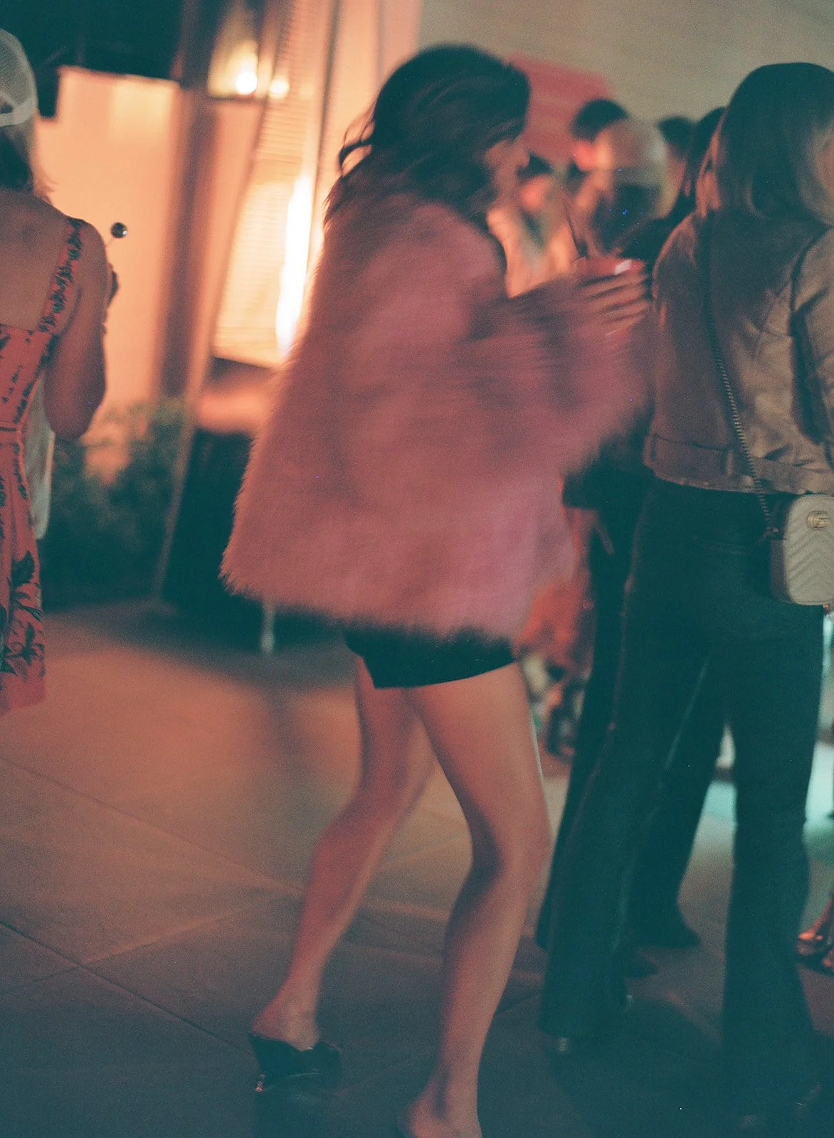 A woman dancing at a party, wearing a short black dress and a pink jacket with fluffy texture, in a dimly lit room with other people.