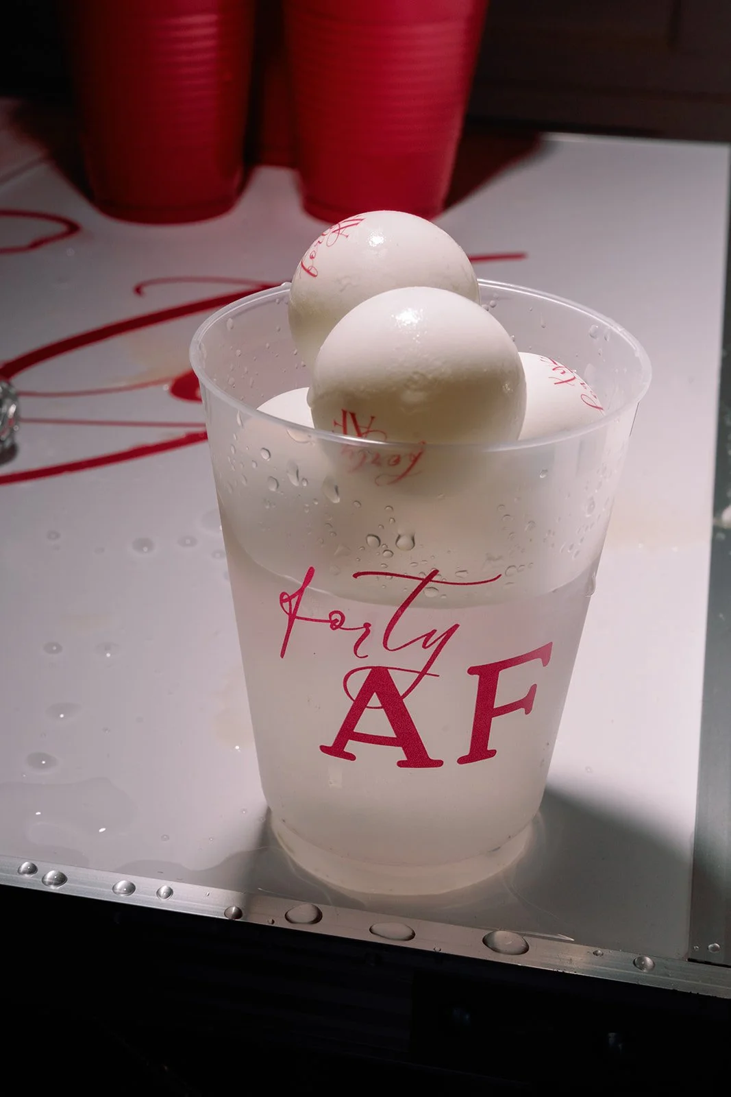A clear cup of water with two white ping pong balls floating inside, labeled 'Forty AF' in red text, with a red backdrop in the background.