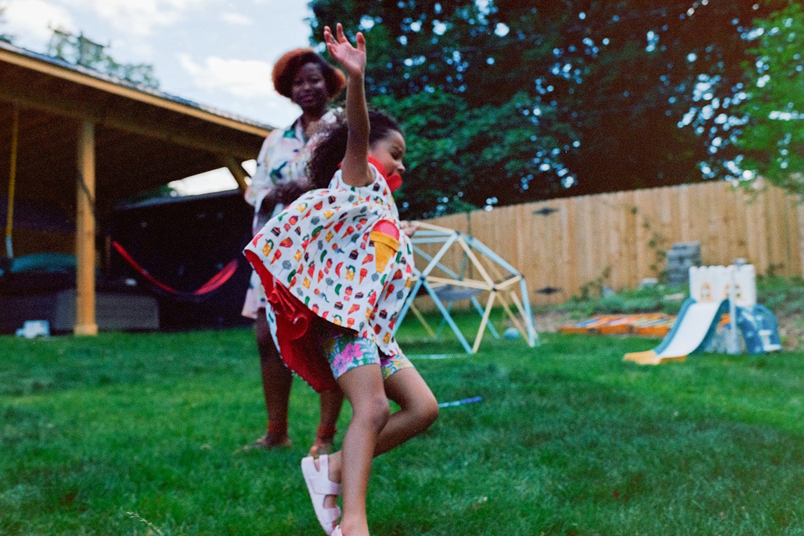 Two girls playing in a backyard, one girl with curly hair is spinning and the other girl with straight hair is waving, in front of a wooden fence and children’s play equipment.