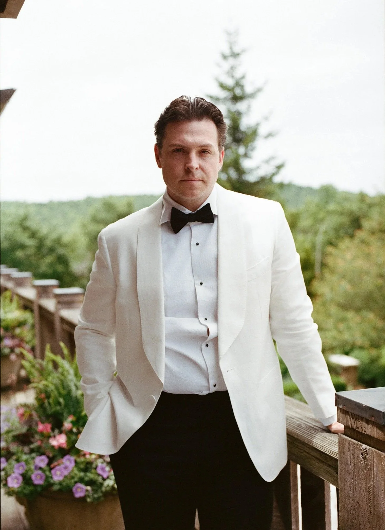 A man dressed in a white tuxedo jacket and black bow tie standing outdoors on a wooden deck with a garden and trees in the background.