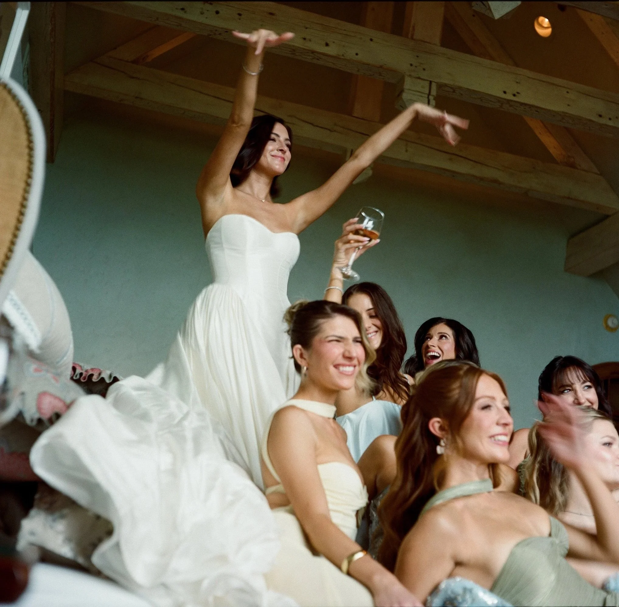 A woman in a white dress holding a glass of champagne and smiling at a celebration party with multiple women seated around her.