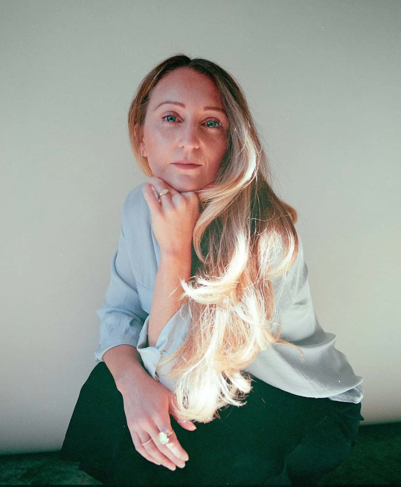 Portrait of a woman with long blonde hair, light blue eyes, and fair skin. She is resting her chin on her hand, wearing a light blue shirt and sitting against a plain, light-colored background.