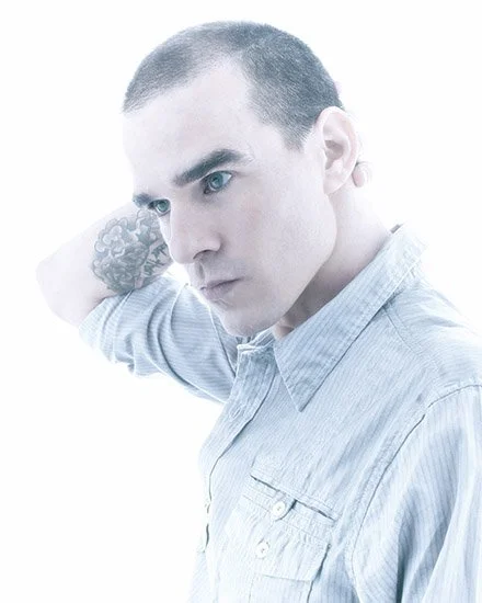Portrait of a man with short dark hair and light skin, wearing a light blue shirt, looking to his left with his right hand behind his neck, and a tattoo visible on his arm.
