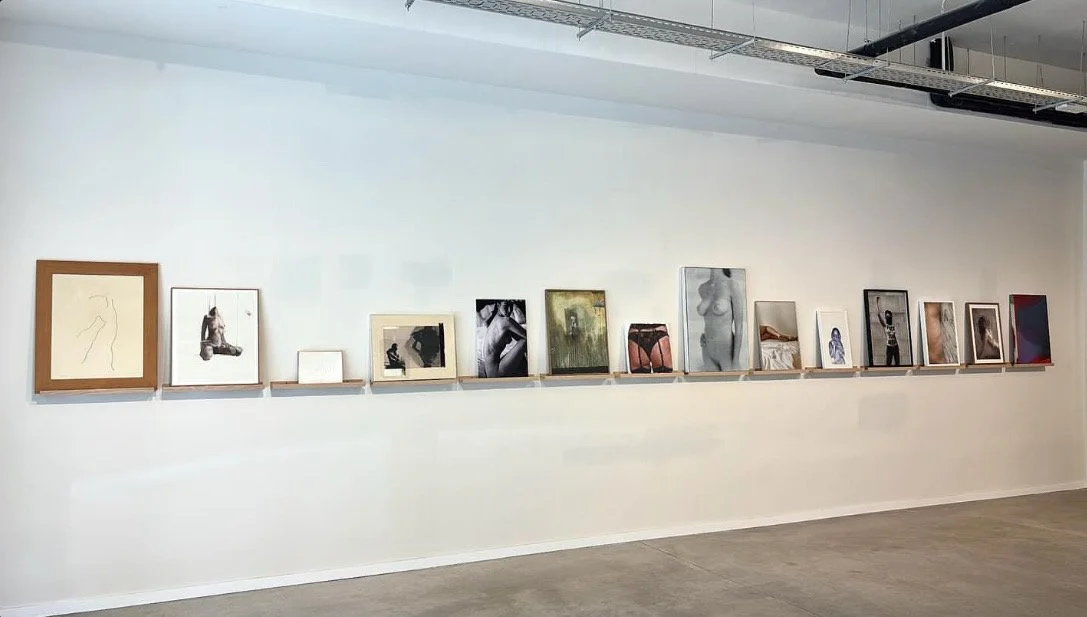 Gallery wall with various framed photographs and artworks displayed on a white wall in an art gallery.A black and white photo of a topless woman sitting on a bed, looking over her shoulder.fine art photograph, Alvaro Alcazar Gallery