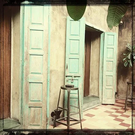 Interior wall with distressed mint green shutters, open to reveal a small nook, with a wooden stool and a potted plant to the right.