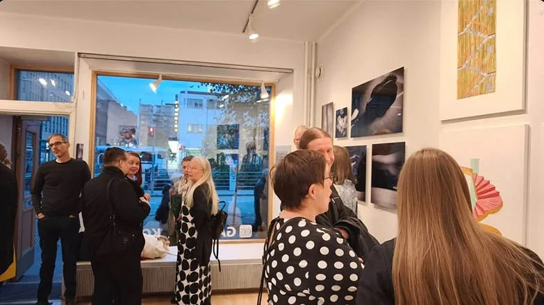 People attending an art gallery exhibition, viewing and discussing various artwork on the walls, with large windows showing city buildings outside.Life Sentence opening exhibition in Helsinki, Finland.