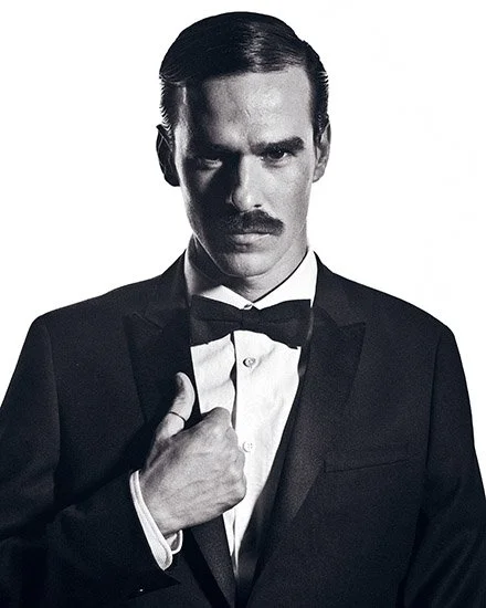 A man in a tuxedo with a bow tie, adjusting his collar, posing with a serious expression in black and white. PHE. Bienal Fundacion Once