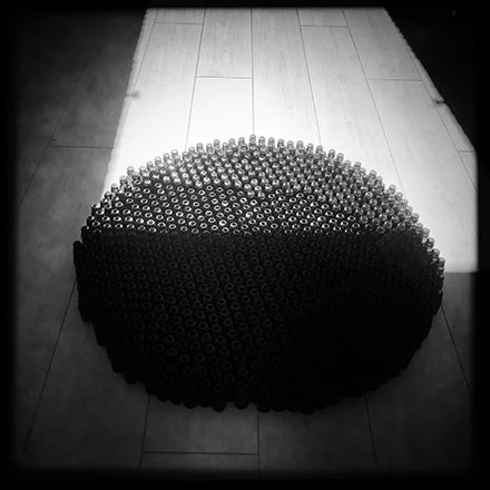 A large collection of black smoke grenades arranged in a semi-circle on a tiled floor, with light coming from a window illuminating the scene.An artist turns her daily dependence on a life-enabling drug into a meditation on survival.