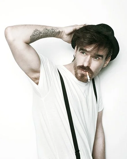 A young man with a beard and mustache, wearing a black hat, white t-shirt, and suspenders, is posing with one arm behind his head, cigarette in his mouth, against a plain white background.PHE. Bienal Fundacion Once